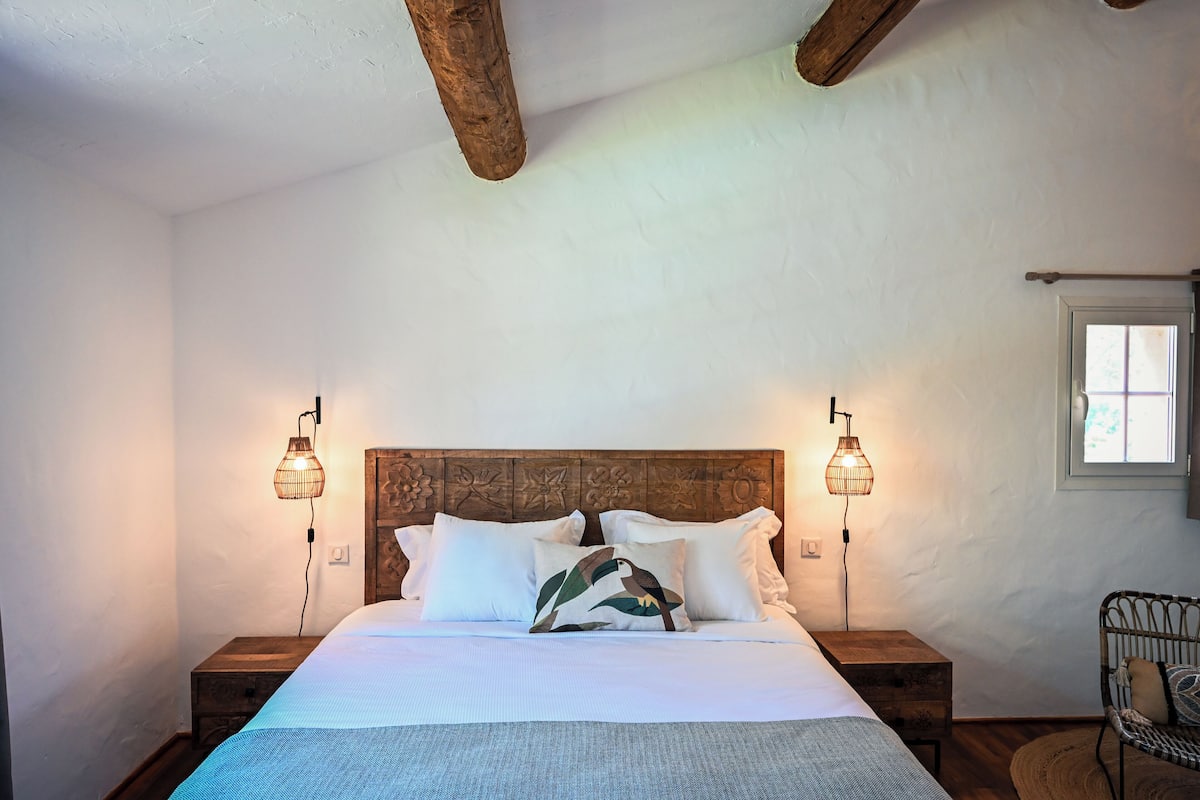 Ein ordentlich bezogenes Bett mit Holz-Kopfteil, zwei Nachttische mit Lampen und ein Korbstuhl.