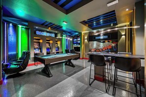 Mesa de bilhar, área de bar e máquinas de jogos num espaço com design moderno e iluminação em tons de verde.
