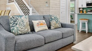 A modern living room with a grey sofa, patterned cushions, a staircase, and a kitchen area with white cabinets. - Coastal Retreat 3 Bedroom Home (Rockport)