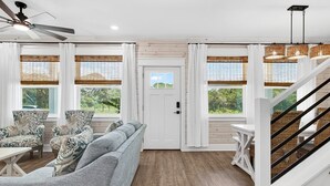 A spacious living room with a grey sofa, patterned armchairs, a wooden floor, and a staircase with wicker railings. - Coastal Retreat 3 Bedroom Home (Rockport)