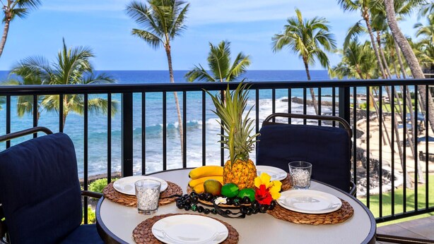 Mesa dispuesta para dos con una decoración de frutas tropicales, copas y platos, con vista a una playa con palmeras y el océano.