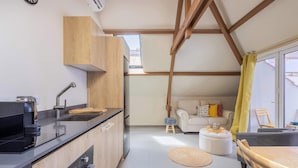 A modern kitchen with a black countertop, wooden cabinets, and a built-in oven. There is a small stool and a white ottoman in the living area with a sofa and a yellow curtain. - Bright Historic Loft in the Heart of Willemstad (Willemstad)