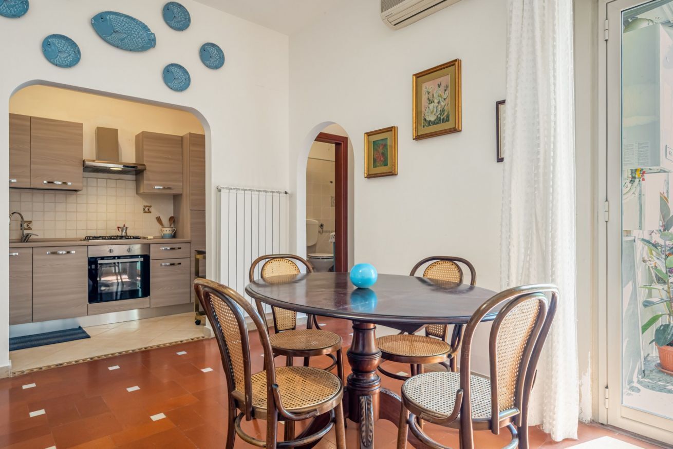 Zona pranzo con tavolo rotondo, sei sedie, un vaso blu e una cucina sullo sfondo.