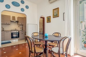 Uma sala de jantar com uma mesa redonda, seis cadeiras, um vaso azul e uma cozinha ao fundo.