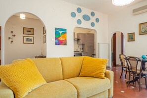 Uma sala de estar com um sofá, uma mesa de jantar com cadeiras e uma cozinha ao fundo.