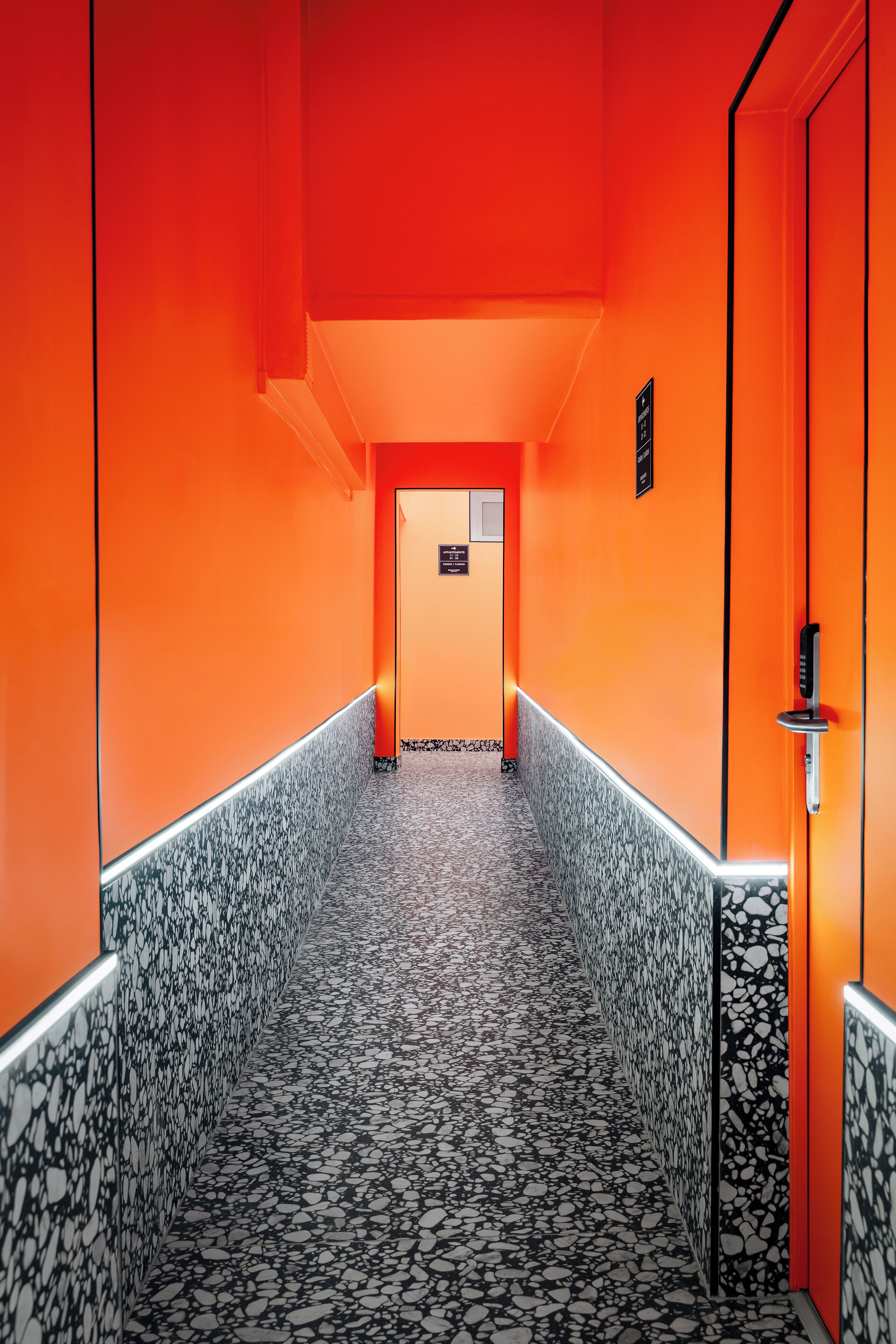 A corridor with a terrazzo floor and orange walls, leading to a door with a keypad lock.