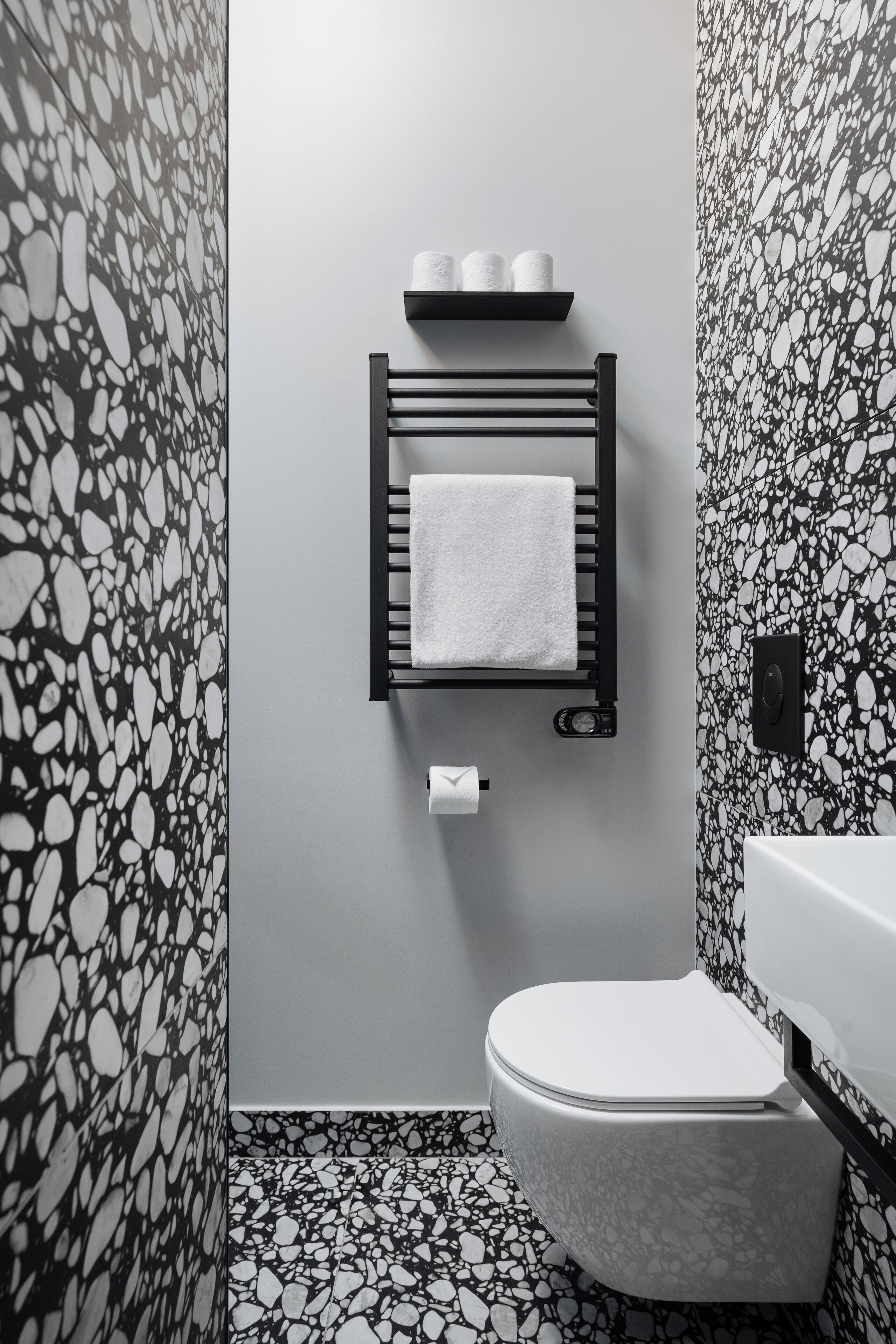A bathroom with a white toilet, a black towel rack with a white towel, and a toilet paper holder. The walls and floor have a black and white speckled pattern.