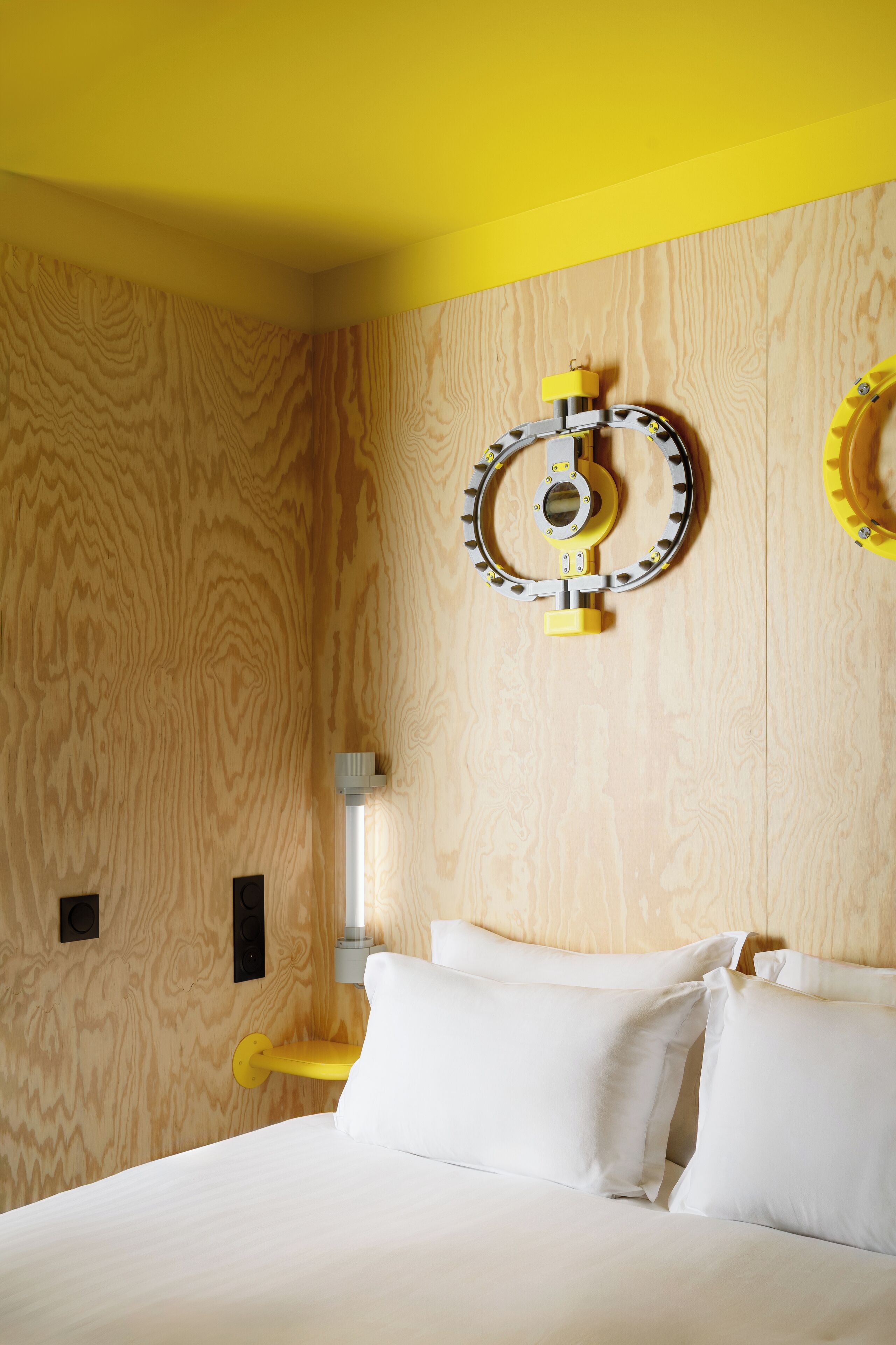 A room with a wooden wall, a bed with white bedding, and a yellow wall-mounted light fixture.