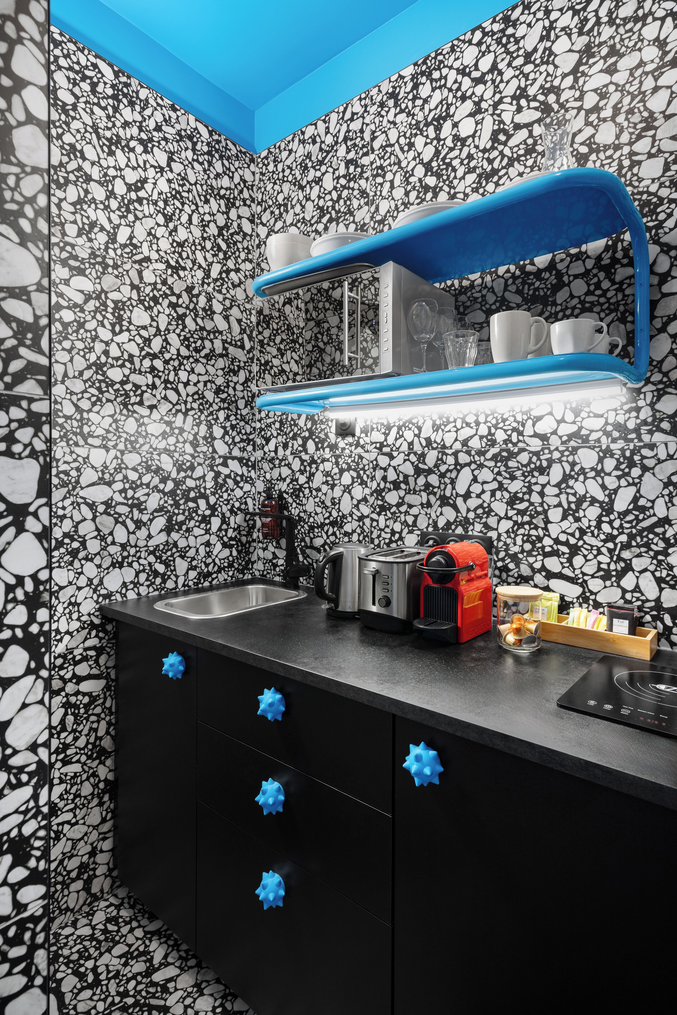 Un baño moderno con salpicadero blanco y negro con motas, un tocador negro con tiradores azules, una cafetera roja y un estante azul sobre el espejo.