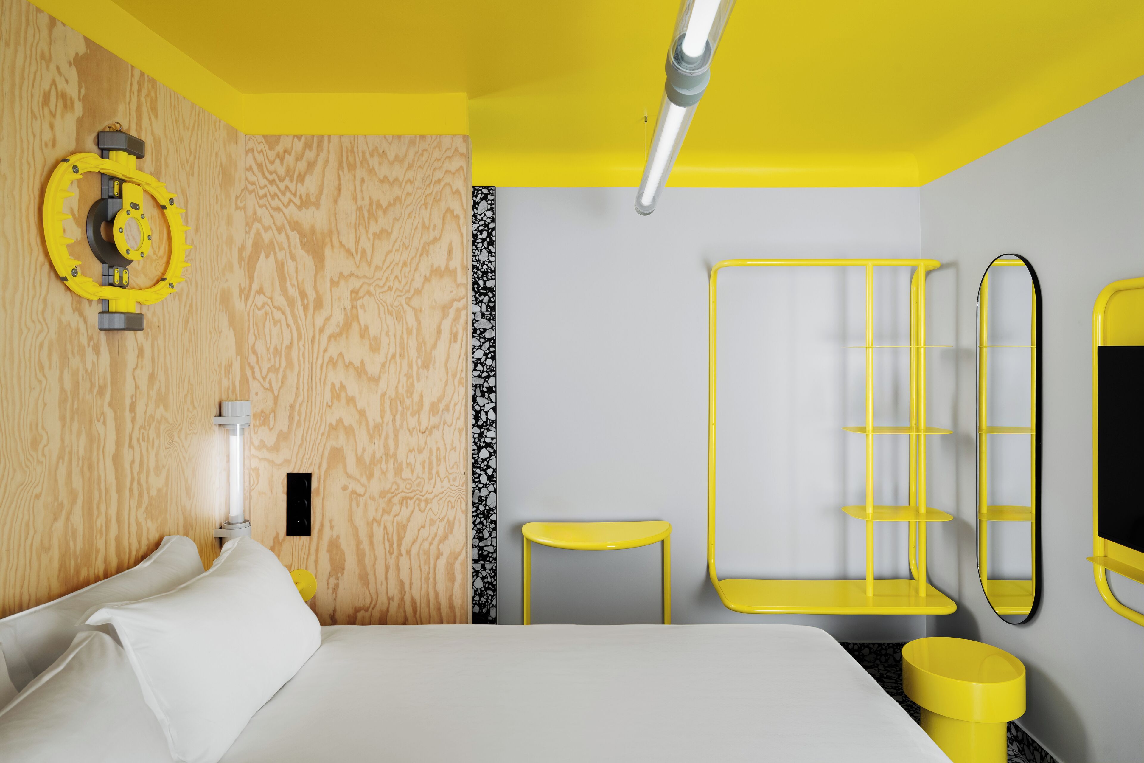 A modern hotel room with a bed, a small table, a yellow sink, and a mirror.