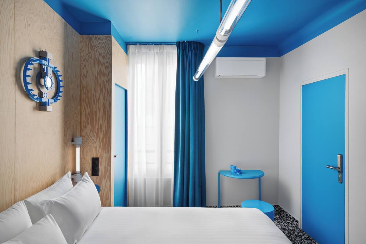 A modern hotel room with a bed, a small table, a blue door, and a curtain.