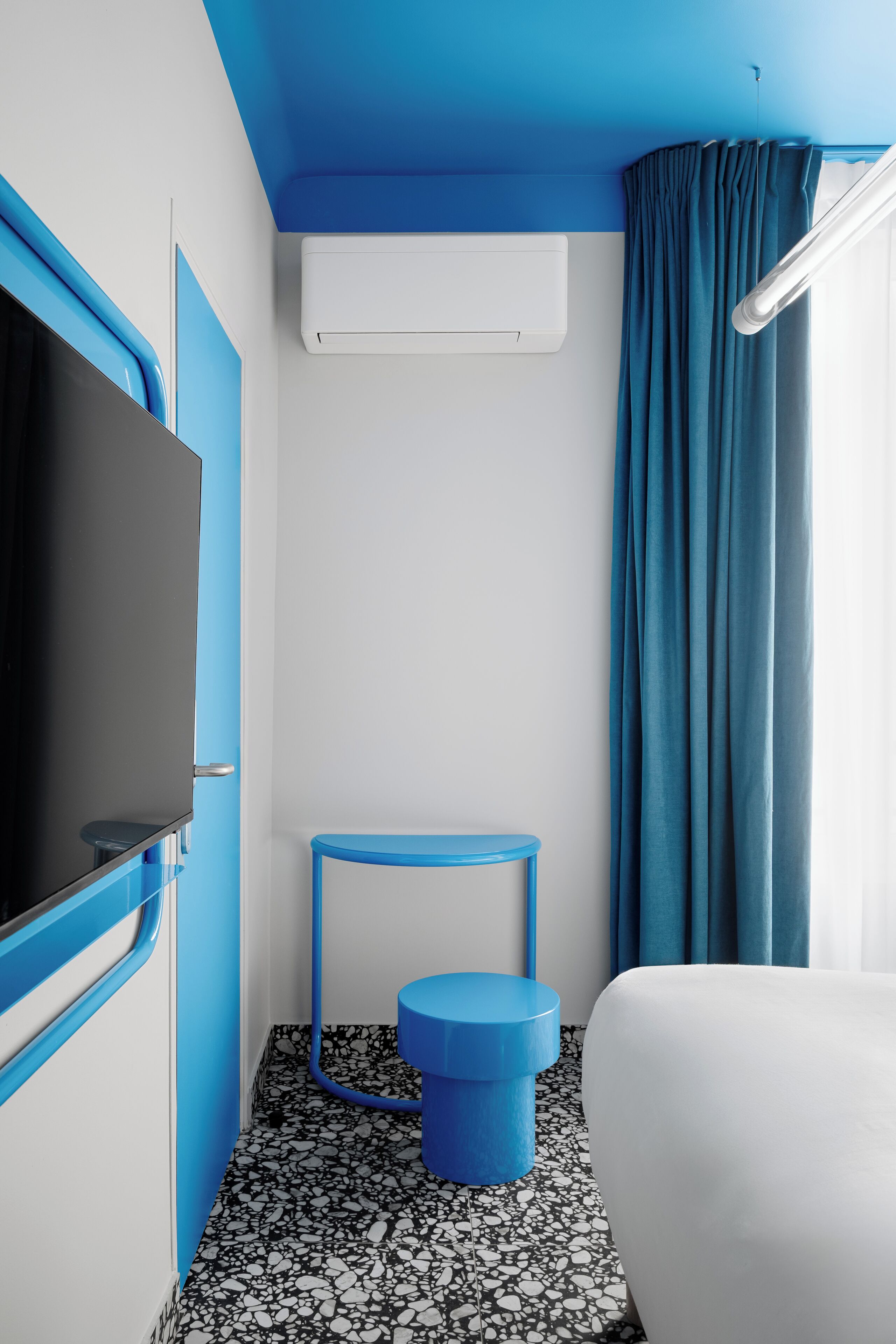 A compact hotel room with a flat-screen TV, a small blue stool, a white bed, and a blue curtain.