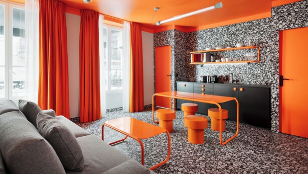 Un salon moderne avec un canapé, une table orange et un coin cuisine doté d’un plan de travail noir et de touches de couleur orange.