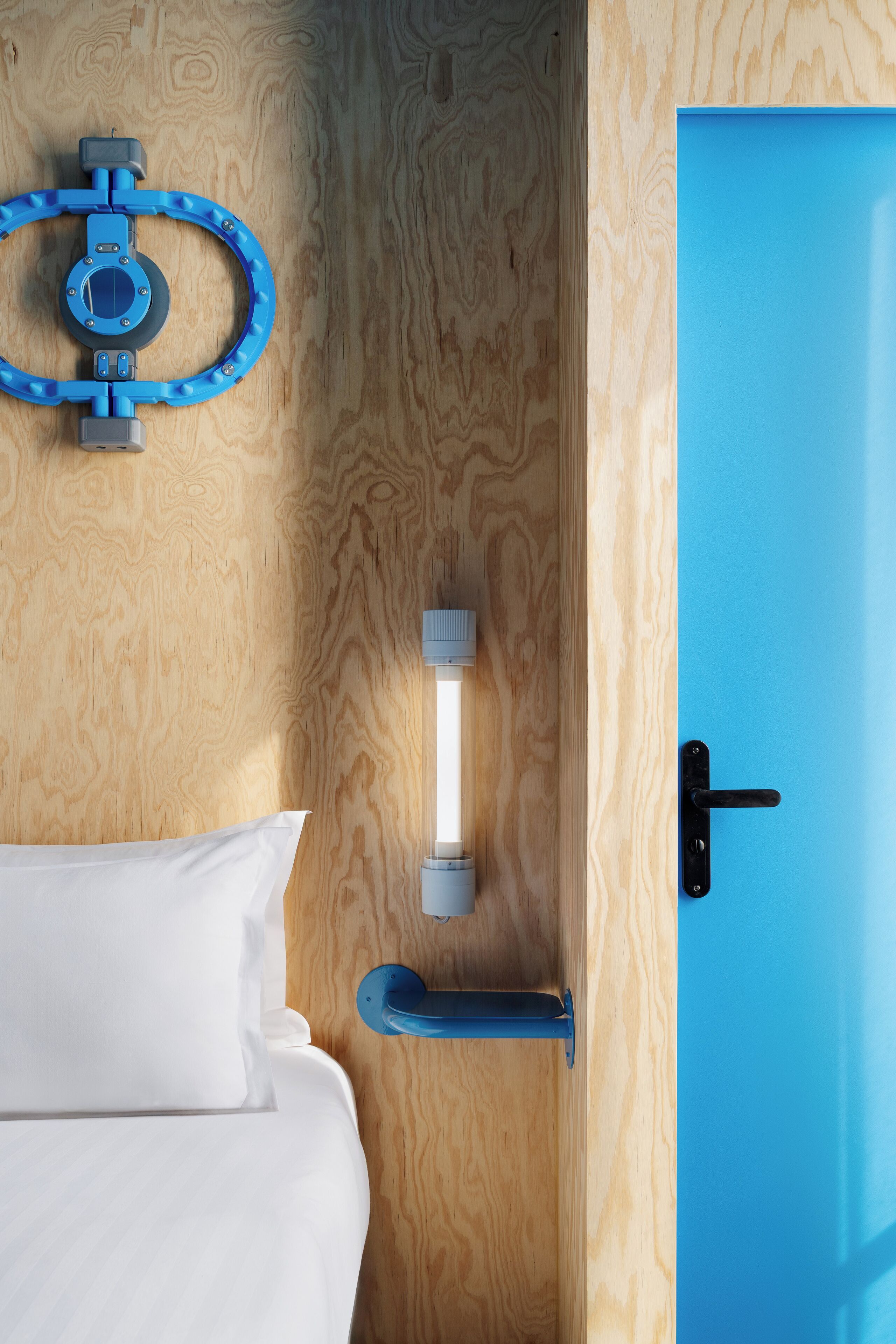 A wooden wall with a blue door, a white bed with a pillow, a blue towel ring, and a wall-mounted light fixture.