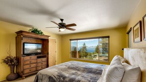 Un dormitorio con cama, ventilador de techo, televisor sobre un mueble de madera, ventana grande con vista a la montaña y una planta en maceta.