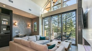 Un salon moderne avec un canapé d’angle, une table basse et un coin repas avec des chaises. Une grande fenêtre offre une vue sur les arbres.