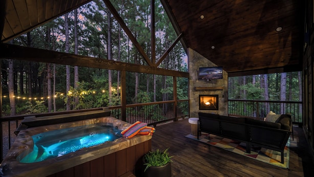 A wooden deck with a hot tub, a stone fireplace, and a couch. - Oakhaven Fell - Couples Retreat - Sauna - Cold Plunge - Hot Tub (Broken Bow)
