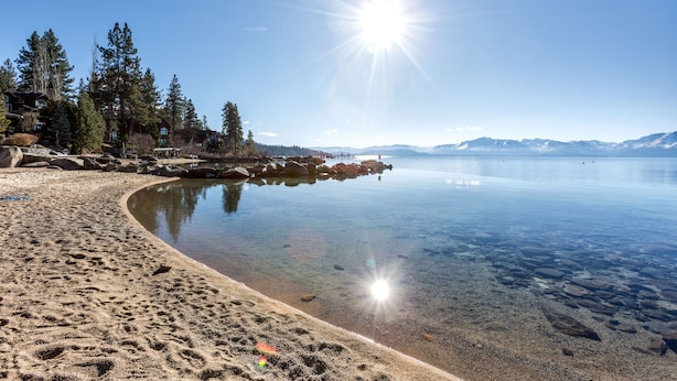 Un lago cristalino con una playa de arena, rodeado de árboles y montañas al fondo.