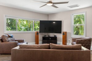 Ein Wohnzimmer mit einem Flachbildfernseher, zwei Lautsprechern, einem Deckenventilator und großen Fenstern mit Blick auf Grünflächen.