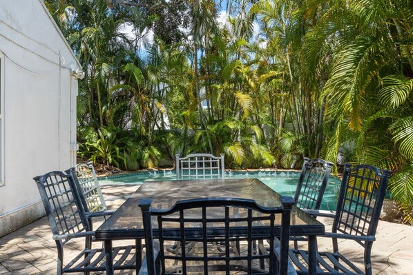 Una mesa con cubierta de vidrio y cuatro sillas, rodeada por una piscina y exuberante follaje tropical.