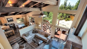 Ein großzügiger Wohnbereich mit einer großen Couch, einem Couchtisch und einem Kamin. Es gibt einen Essbereich mit Tisch und Stühlen, und im Hintergrund eine Küche. Große Fenster bieten Blick ins Freie.
