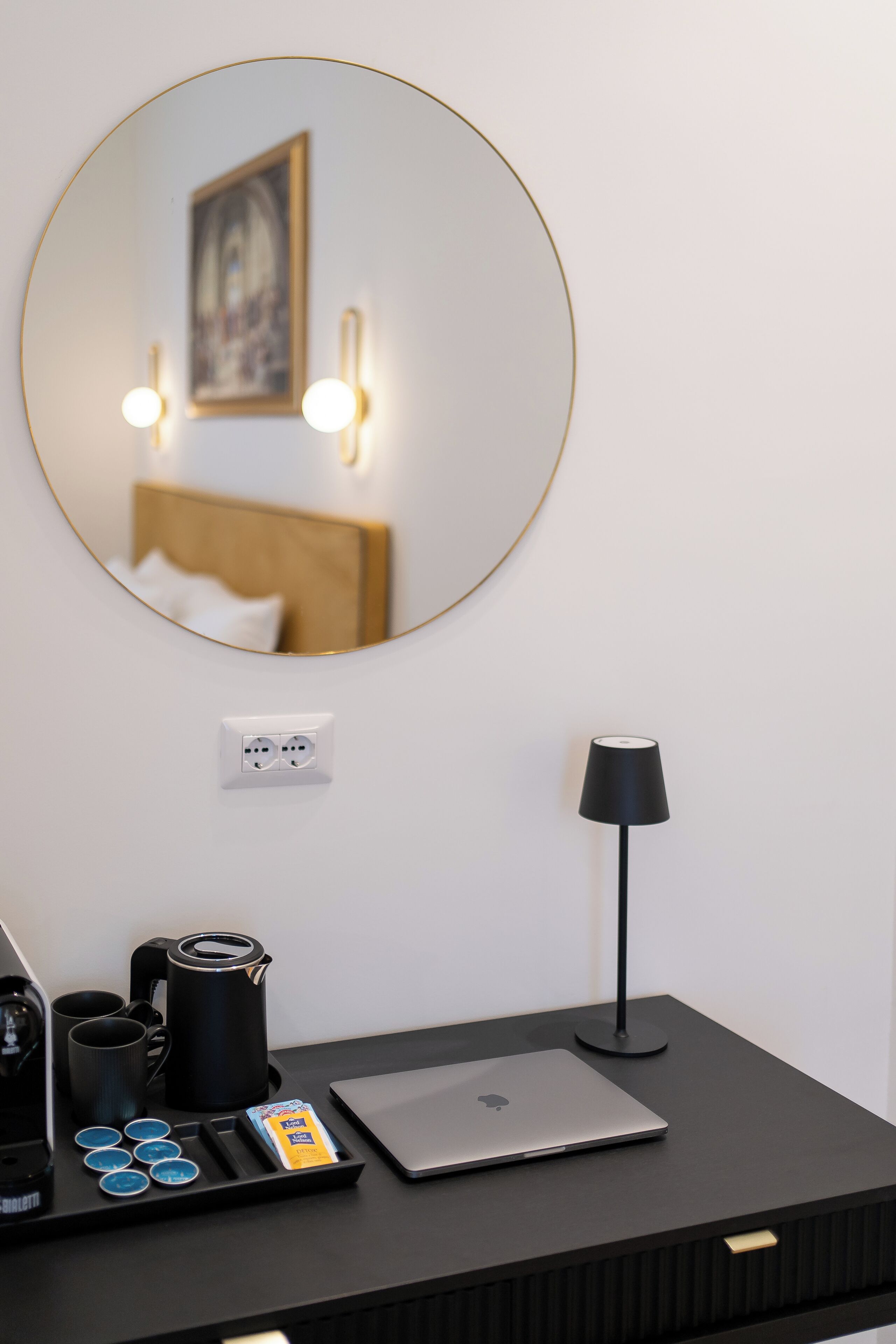 Une chambre d’hôtel avec un miroir rond, un lit avec une literie blanche, une table de chevet noire avec une lampe, un ordinateur portable et un coin café.