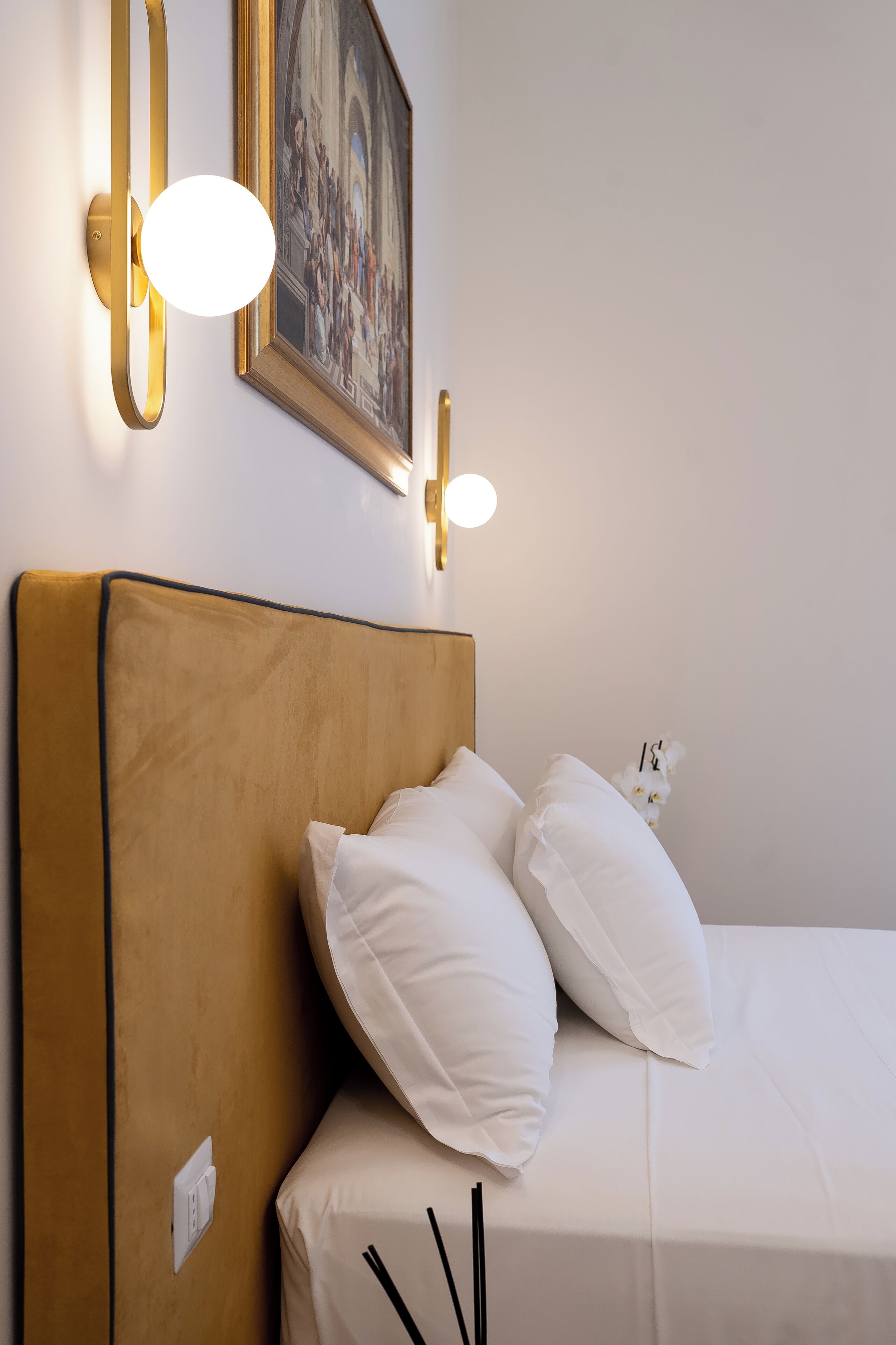 A hotel room with a single bed, a headboard, two wall-mounted lights, and a framed painting above the bed.