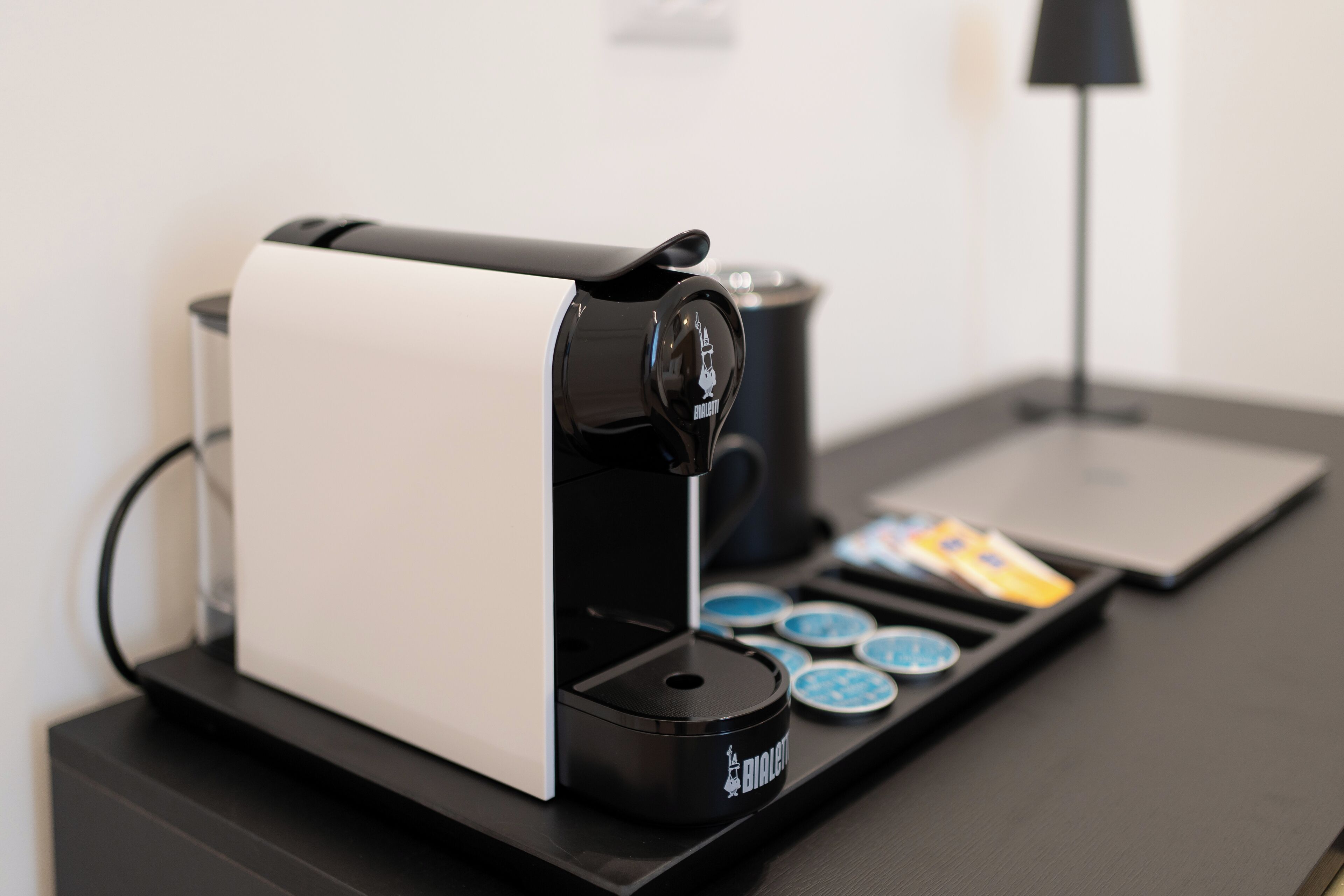 Una cafetera con diseño blanco y negro, un porta cápsulas de café y un ordenador portátil sobre un escritorio.
