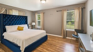 A bedroom with a tufted headboard, a bed with white linens, a desk with a chair, and a large window with curtains. - Cozy Temple Home Fenced Yard & Wi-fi 3 Bedroom Home (Temple)