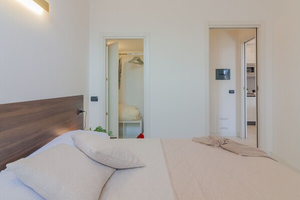 A bedroom with a wooden headboard, a bed with beige bedding, and a small bedside table with a plant. There are two doors leading to other rooms.