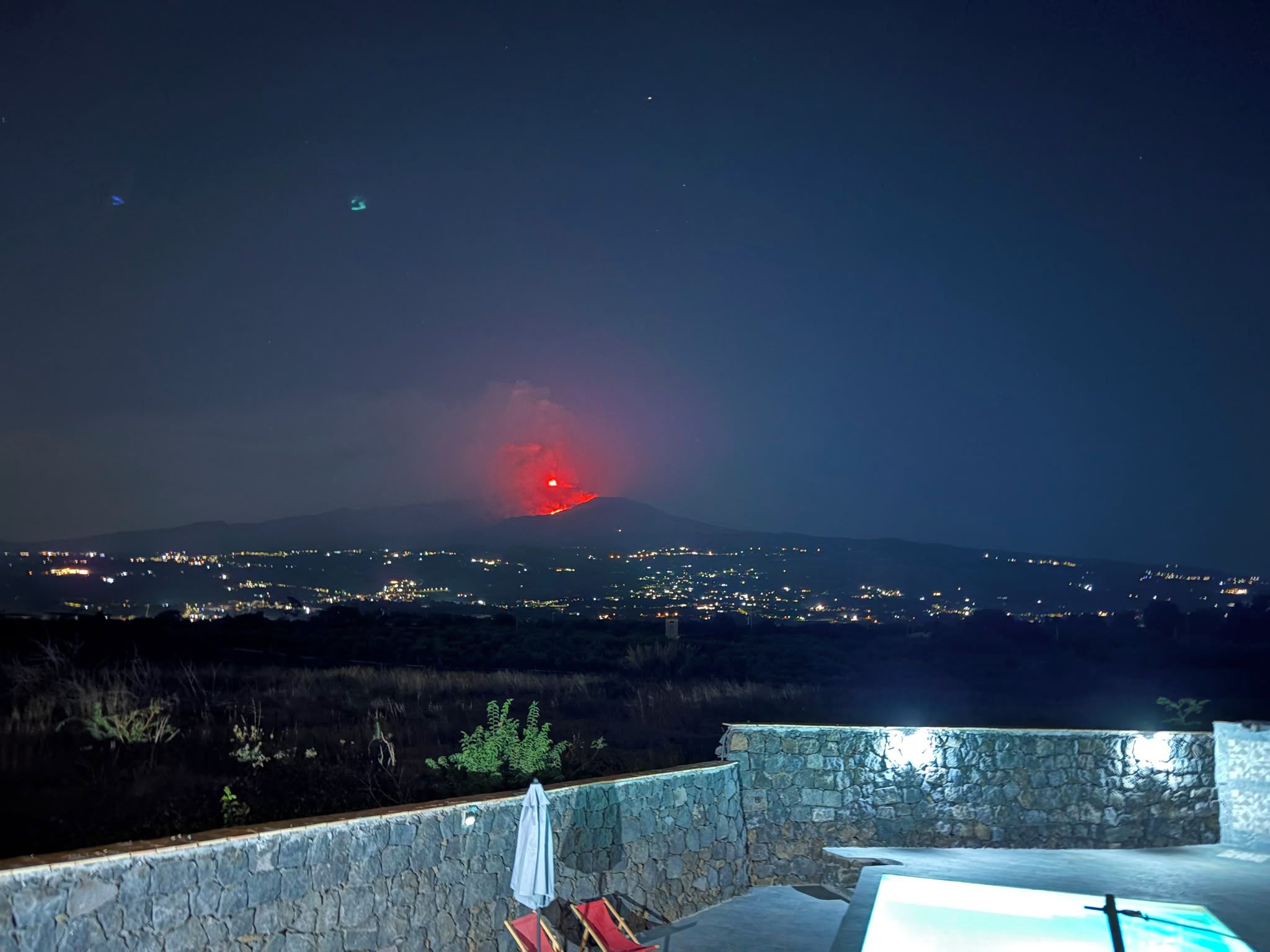 Un muro di pietra con piscina e sedie a sdraio, con vista su una città e una montagna illuminata all'orizzonte.