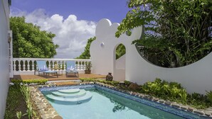 Una piscina con borde de piedra y filo de azulejos, rodeada de follaje y una pared blanca con un recorte circular.
