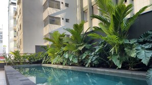 A swimming pool with a tiled edge, surrounded by lush green plants and a building with multiple windows. - Setin Pamplona R (Sao Paulo)