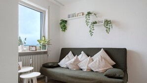 Een kamer met een groene bank, witte kussens, een klein witte tafel en een raam met planten.