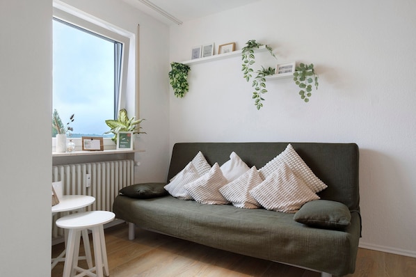 A room with a green sofa, white cushions, a small white table, and a window with plants.