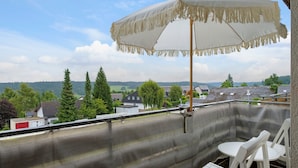 Een balkon met een witte parasol, een witte stoel en uitzicht op huizen en bomen.