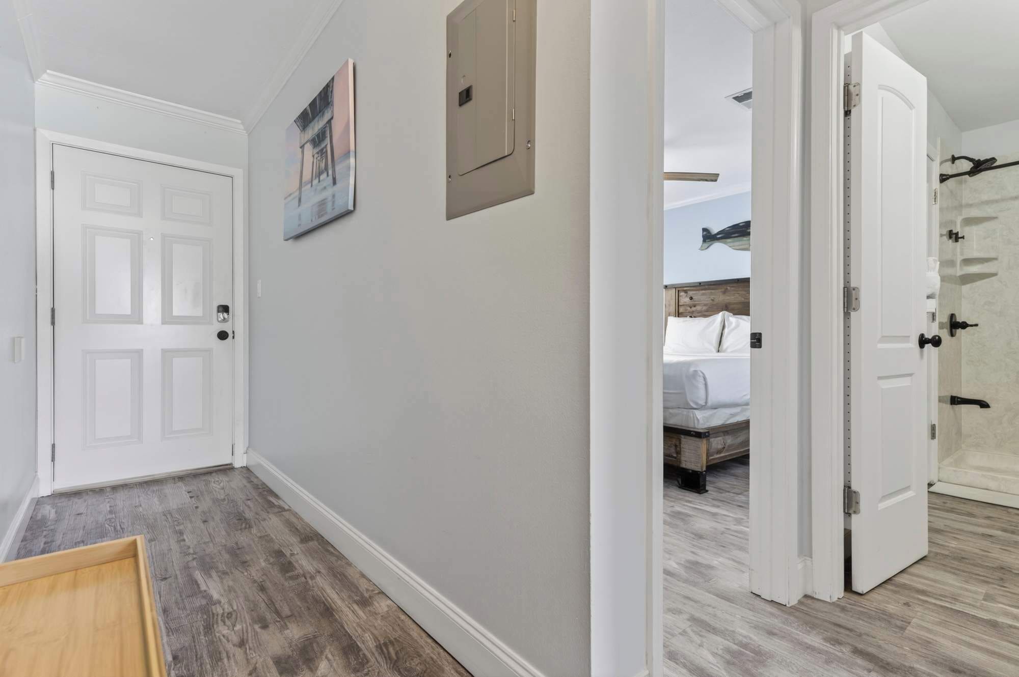 Un couloir avec une porte blanche, un tableau encadré au mur, et une vue sur une chambre à coucher avec un lit et une tête de lit en bois.
