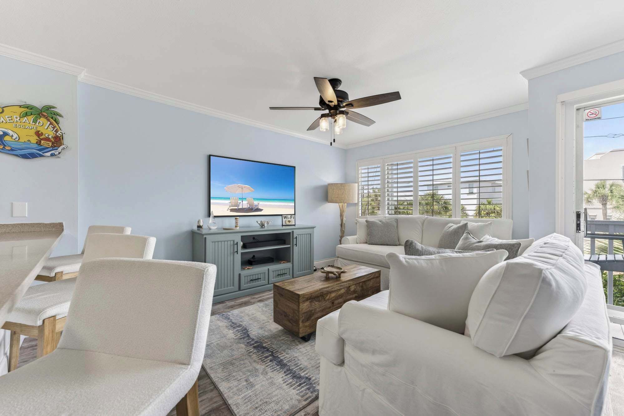 A living room with a TV, white furniture, a ceiling fan, and a view of the outdoors.