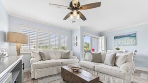 Ein Wohnzimmer mit zwei weißen Sofas, einem Holztisch, einem Deckenventilator und einem Gemälde mit Strandblick.