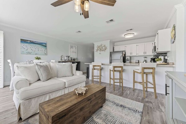A modern living room with a white sofa, wooden coffee table, and a kitchen area with white cabinets and a bar stool.