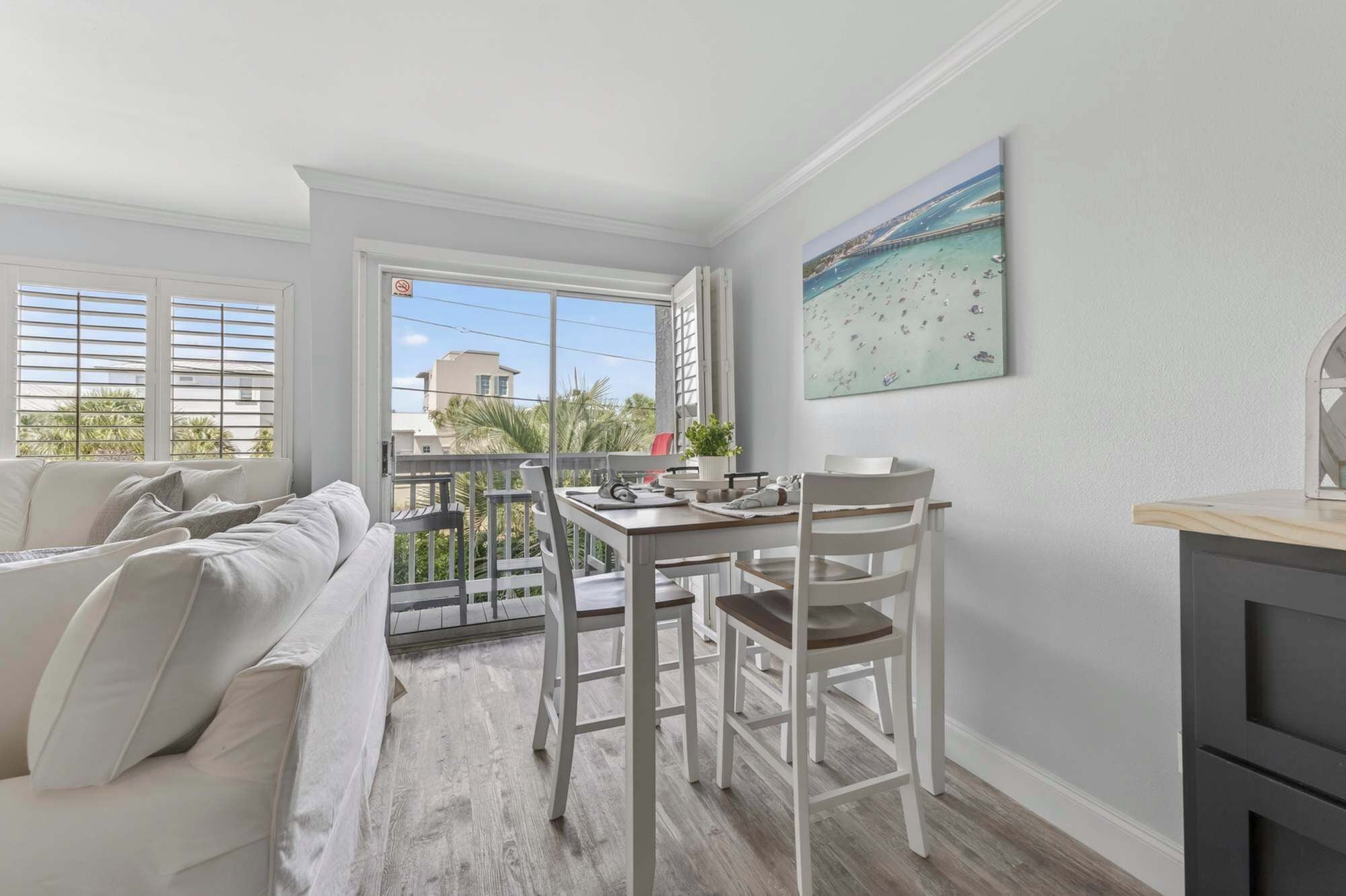 A balcony with a view of palm trees and buildings, a white sofa, a dining table with chairs, and a painting of a beach scene.
