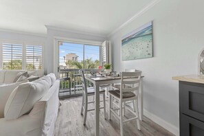 A balcony with a view of palm trees and buildings, a white sofa, a dining table with chairs, and a painting of a beach scene. - Holiday Isle - Private Beach - Special Offer! (Destin)