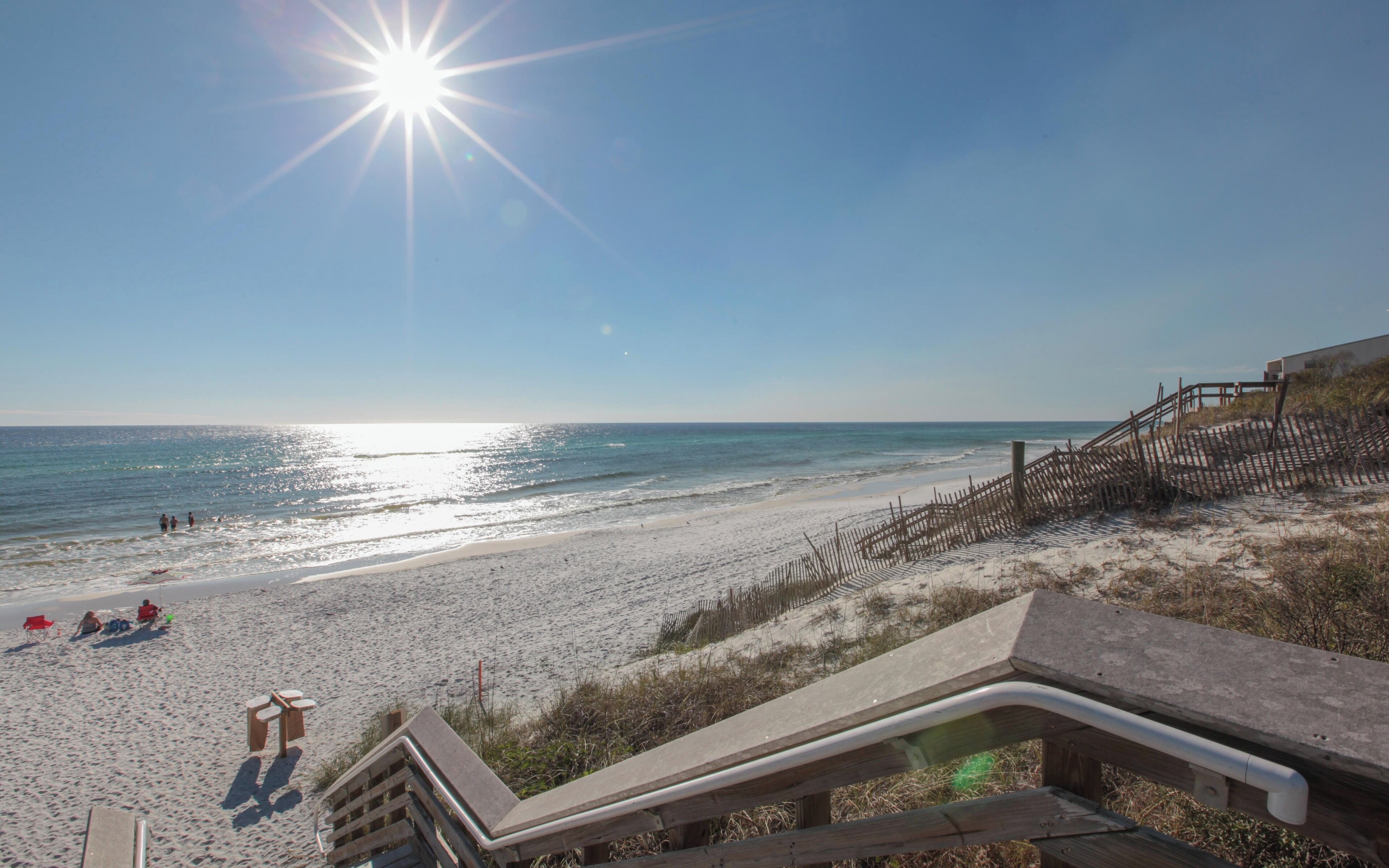 A beach with a wooden staircase leading down to the sand, a clear sky, and the ocean in the background.