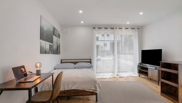Un dormitorio con cama, escritorio con ordenador portátil, silla, televisor y ventana con cortinas.