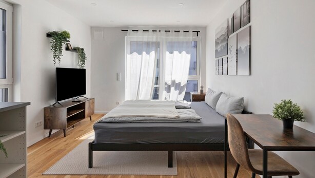 Un dormitorio moderno con cama, mesita de noche, televisor y ventana con cortinas.