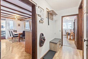 Una habitación con piso de madera, una mesa de comedor con sillas, un banco, un zapatero en la pared y una puerta que da a otra habitación.