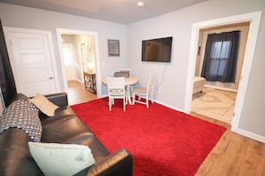 A living room with a red carpet, a brown leather sofa, a white dining table with chairs, a flat-screen TV mounted on the wall, and a window with curtains. - Spacious 5 BR Stay in Westminster | Perfect for Groups & Longer Stays (Westminster)
