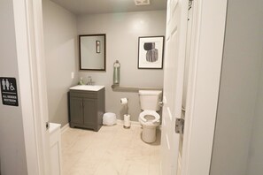 Une salle de bain équipée de toilettes, d’un lavabo et d’un meuble de rangement, ainsi qu’un panneau indiquant qu’il s’agit de toilettes à usage mixte, et deux tableaux encadrés accrochés au mur.