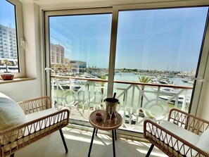 Een balkon met rieten meubilair, een tafeltje met een schaal en glazen, en een uitzicht op een jachthaven met boten en gebouwen.
