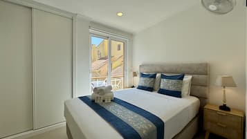 Habitación de hotel con cama, mesita de noche, lámpara y ventana con vistas a edificios.